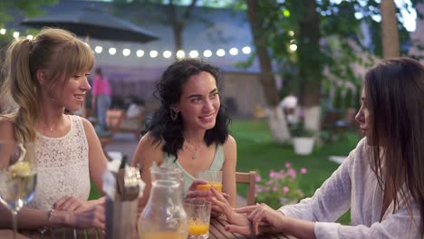 Three-european-women-sit-in-the-outdoors-cafe,-drink-juice-and-have-fun-communicating