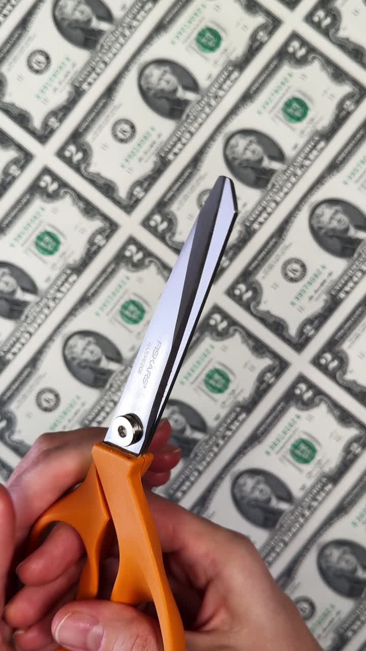 San Antonio, USA, 14 August 2025: Female hands open the scissors with orange handles. Person is going to cut the sheet of two dollar banknotes lying at backdrop. Vertical video