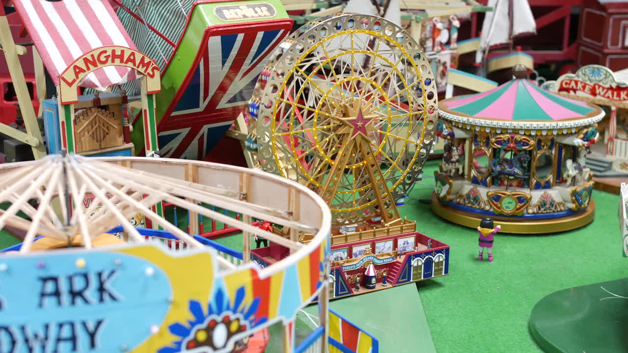 A model ferris wheel with a gold star center is surrounded by fairground structures, including a helter skelter, carousel, and figurines on a green platform