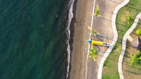 Vista-Aérea-De-Arriba-Hacia-Abajo-Del-Barco-Jukung-Indonesio-Varado-En-La-Hermosa-Costa-De-Lovina-Bali-Indonesia-Mientras-Las-Olas-Azules-Del-Océano-Chocan-Al-Atardecer