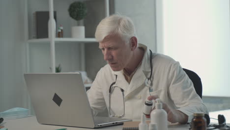 a grey haired, middle aged doctor consults on his laptop the composition of a medicine