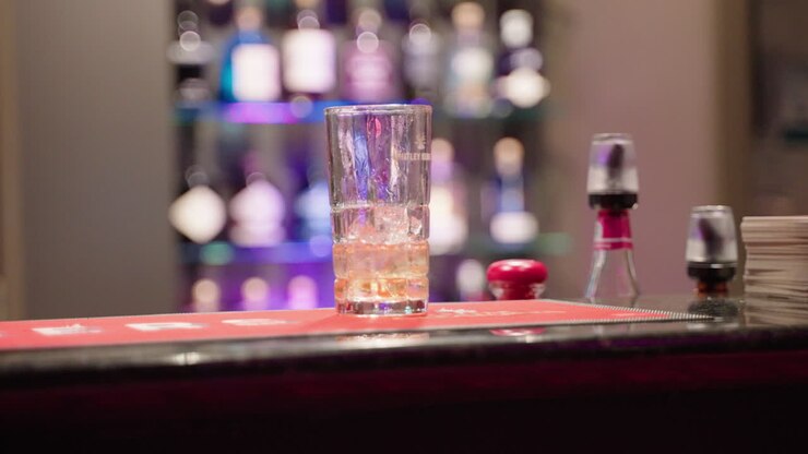 Panoramica 4K al rallentatore di un bicchiere di bevanda ghiacciata poggiato sul bancone del bar davanti a un espositore di alcolici
