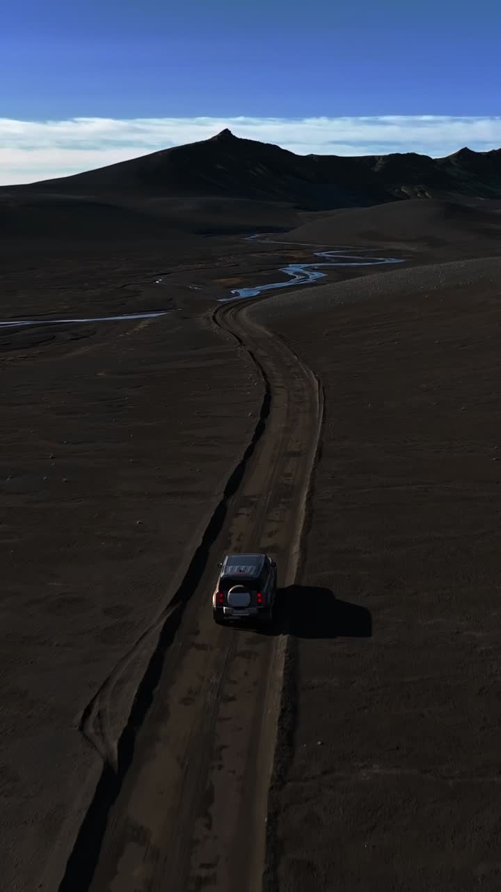 Inseguimento aereo di un SUV che attraversa scuri altopiani vulcanici su una pista sterrata tortuosa verso corsi d'acqua intrecciati e cime distanti, creando una drammatica scena di viaggio per spedizioni remote.