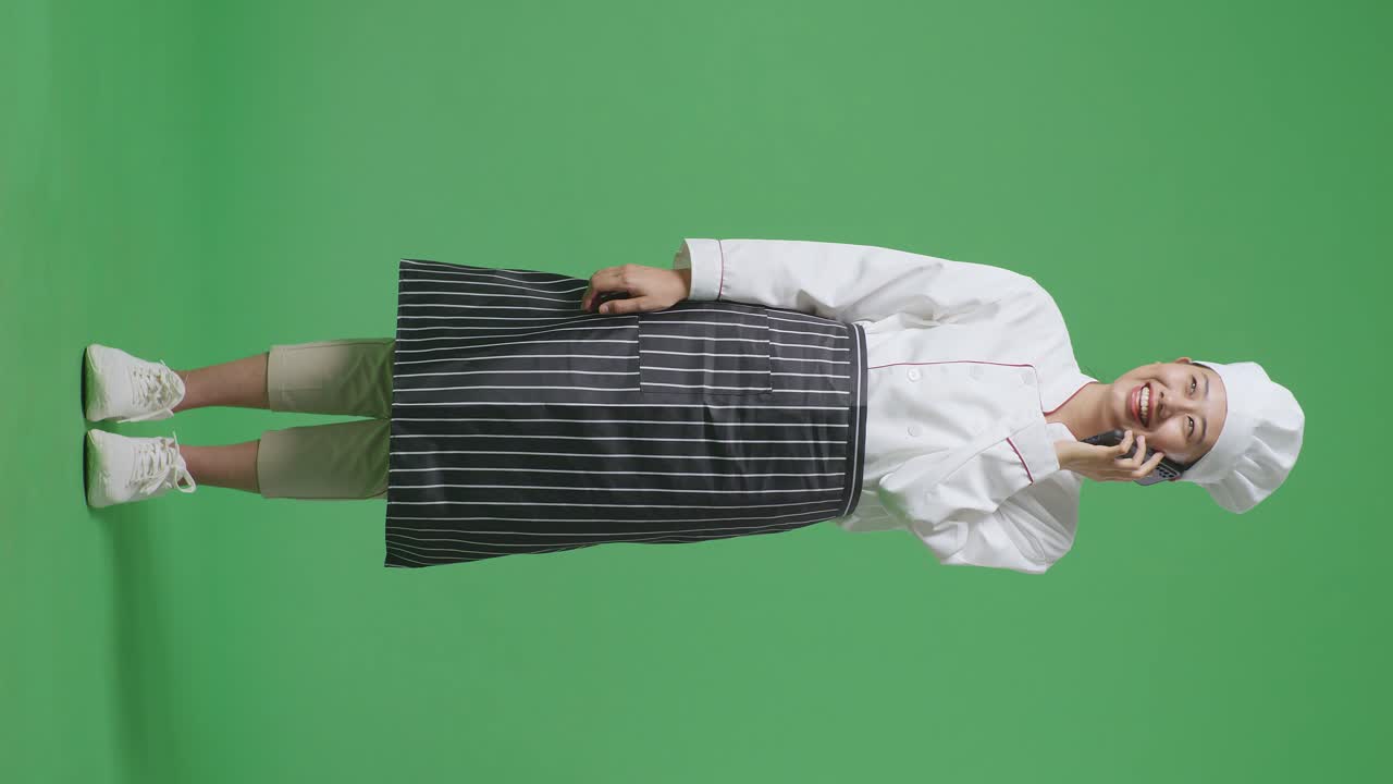 Full Body Of Asian Woman Chef Talking On Smartphone And Looking Around While Standing In The Green Screen Background Studio