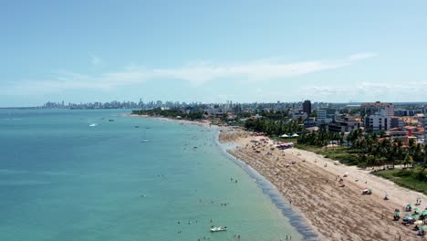 Toma-Aérea-Descendente-De-La-Playa-De-Bessa-Durante-La-Marea-Baja-En-La-Capital-Tropical-De-Joao-Pessoa,-Paraiba,-Brasil-Con-Gente-Disfrutando-Del-Océano-Y-Barcos-De-Pesca-En-La-Costa-En-Un-Día-De-Verano