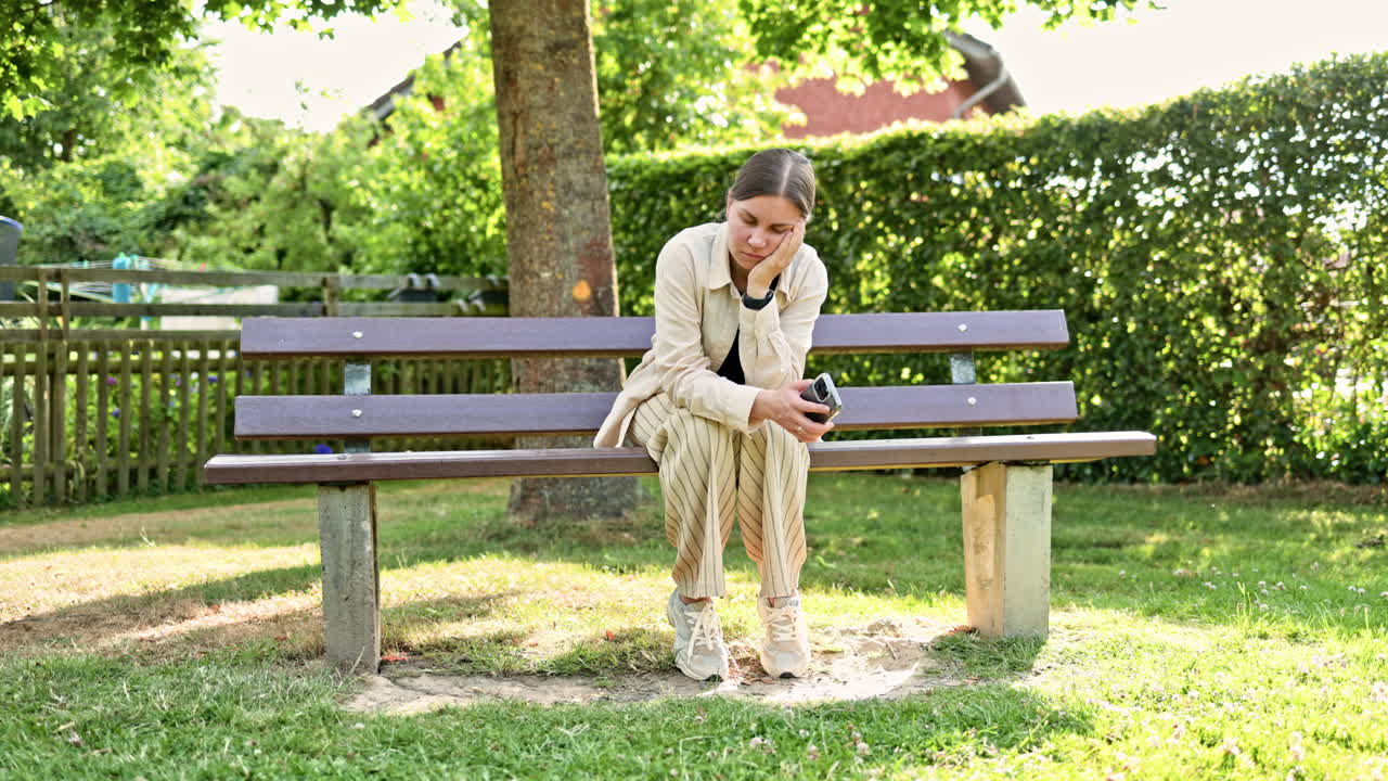Woman with phone sitting in park on bench and enjoying mobile conversation on sunny morning. Smiling girl talking on mobile phone surrounded by greenery. Happy woman enjoying talking on phone