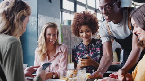 four female friends take photos of food in restaurant to post on social media as waiter serves meals