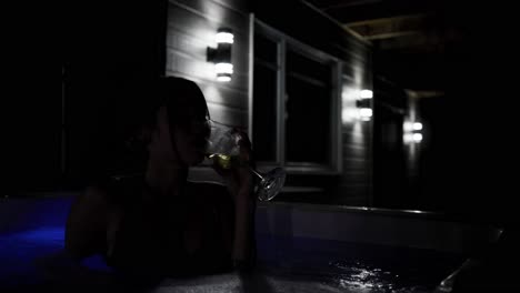 young woman drinking a glass of wine while relaxing in a hot tub outside a cozy cabin in the evening