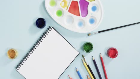 overhead view of notebook, brushes and paints on blue background, in slow motion