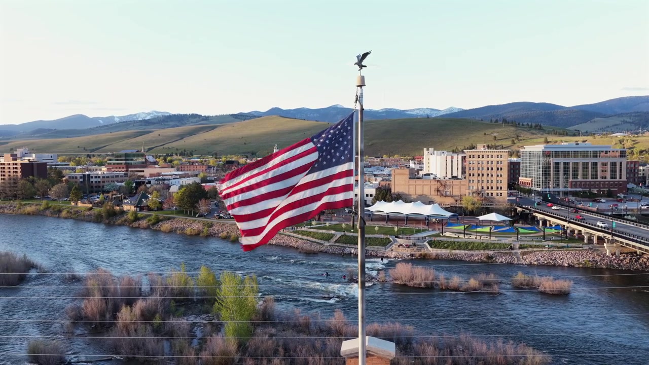 Suuri Amerikan lippu tangossa, josta avautuu näkymä Missoulan keskustaan, Clark Fork -joelle koskineen, maantiesillalle, jokivarsipuistoon ja lumihuippuisille vuorille kirkkaan sinisen taivaan alla. Montana, USA. Laaja kiertävä ilmakuva