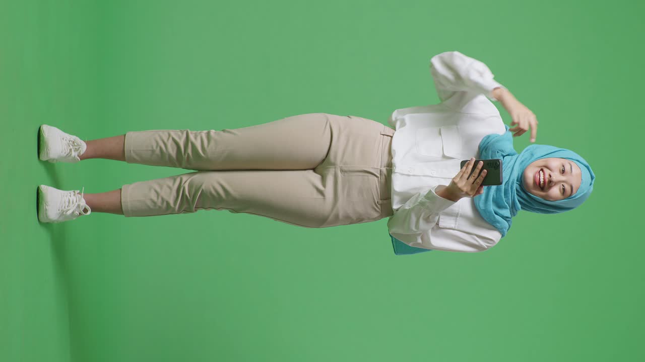 녹색 스크린 배경 스튜디오에 서 있는 동안 손에 스마트폰을 가리키며 미소 짓는 아시아 무슬림 여성의 몸이 가득 차 있습니다.