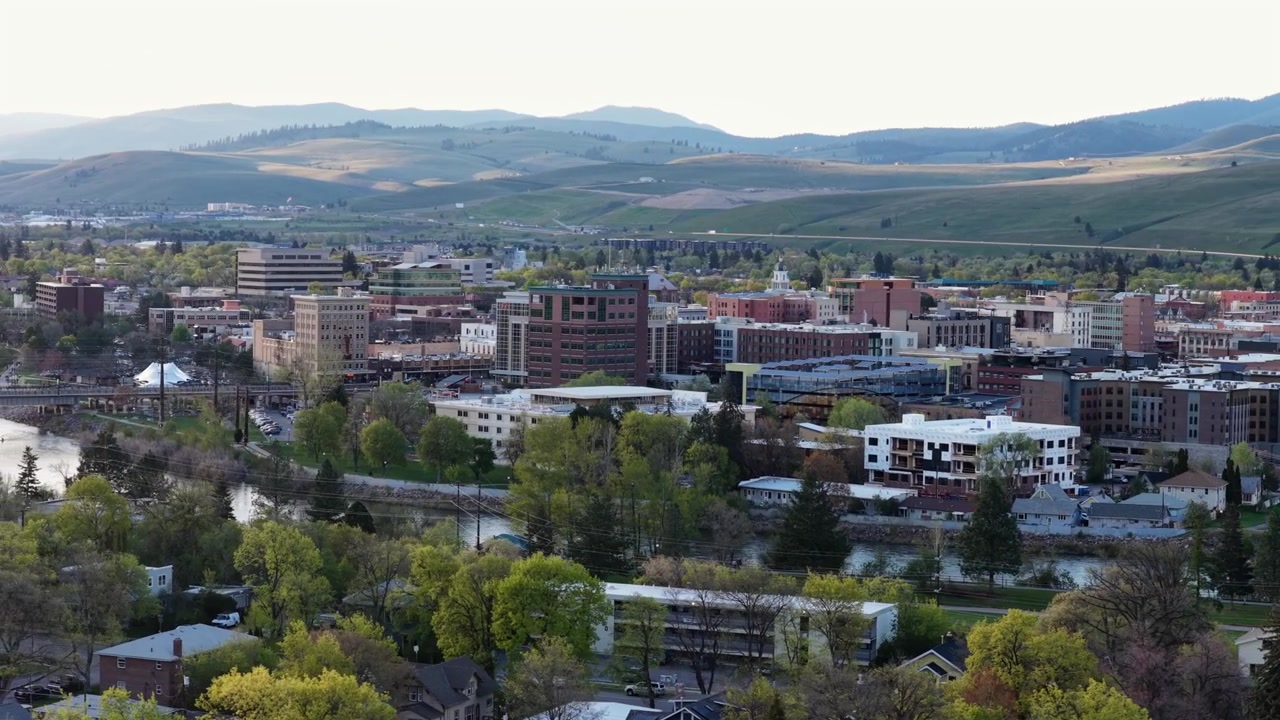 Missoula sentrum med mursteinsbygninger, Clark Fork River, trebevokste boliggater, hjem og bølgende grønne åser med fjellkjede under en myk kveldshimmel. Montana, USA. Vidvinkel luftfoto