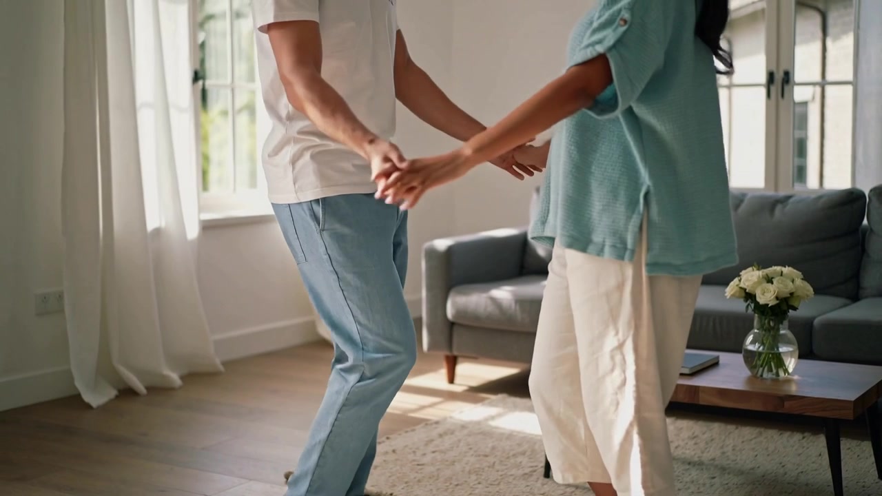 Dancing white-tee man and teal-top woman holding hands in living room near window, sunlight drawing