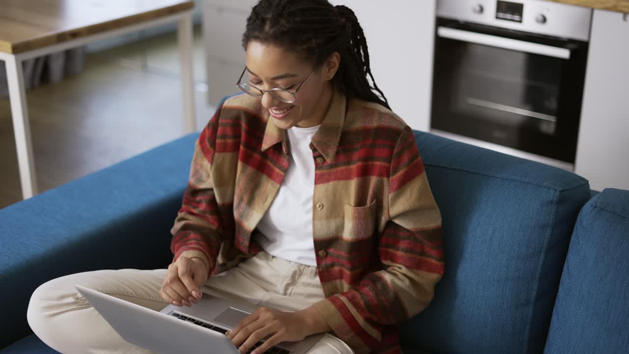 Positive girl with dreadlocks is typing on laptop on couch, laughing and smiling
