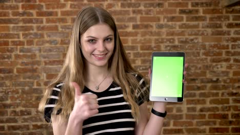 young happy blonda is showing green screen of tablet, like sign, communication concept, brick background