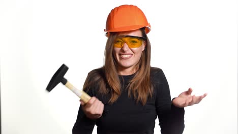 a beautiful woman in orange eyeglasses and helmet is holding a hammer in her hands and hitting herself by it in her hand on a white background in the studio. engineer