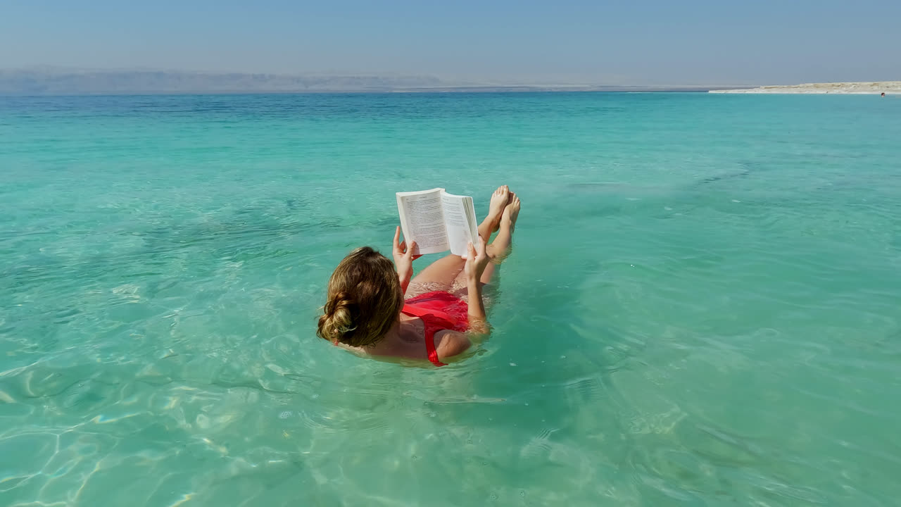 Beautiful Young Woman Floats in the Dead Sea in Red Swimsuit while Reading Book
