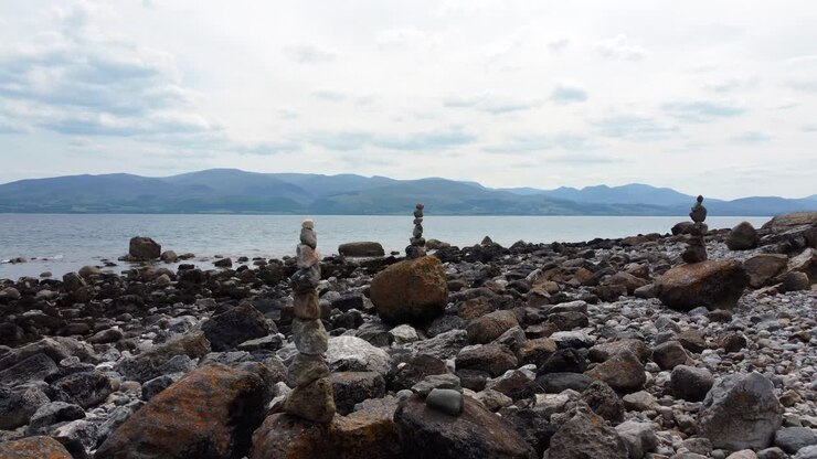 Ciottoli da meditazione per la spiritualità equilibrata impilati su una costa di spiaggia rocciosa e aliena con catena montuosa, dolly a sinistra.