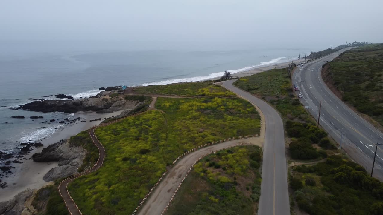 Waves roll into a rocky beach on the Southern California coast as the fog obscures the ocean. A highway can be seen on the right of the shot, contrasting the lush scenery and oceanscape.