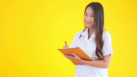 asian nurse doctor woman writing notes in the clipboard on yellow isolated background smiling