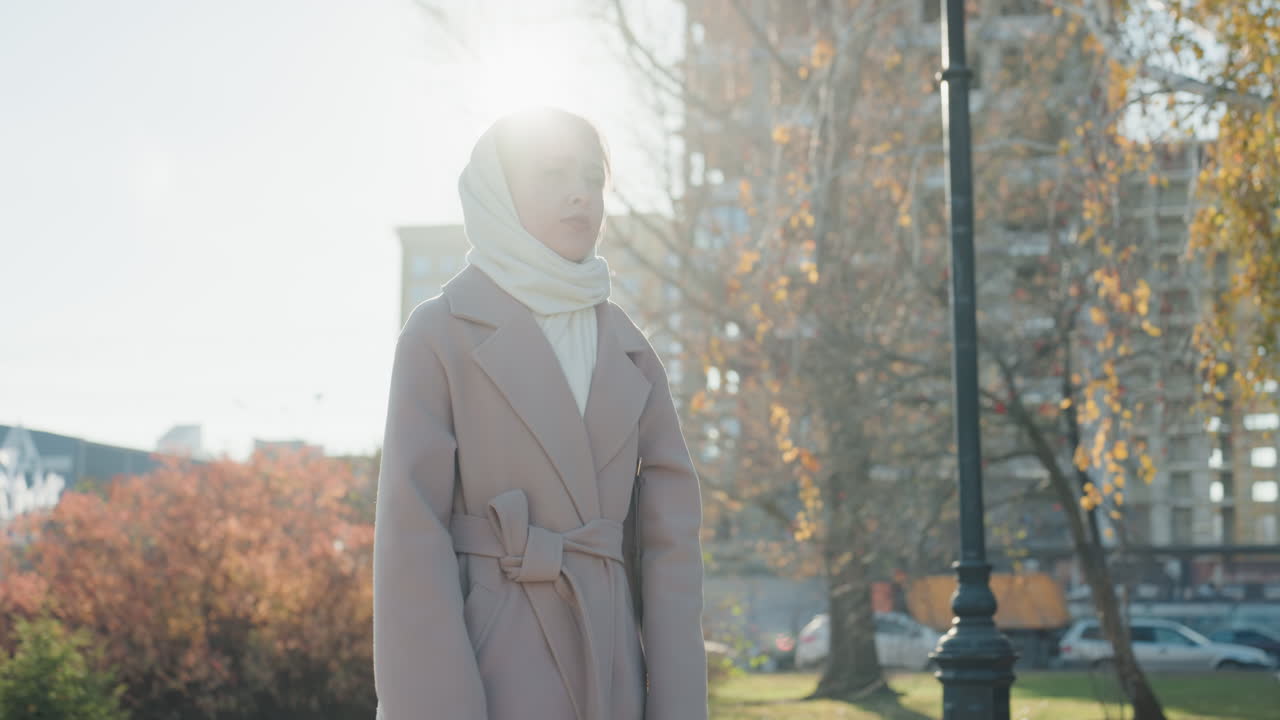 Giovane donna in sciarpa, parco autunnale. Morbida luce in controluce illumina il cappotto beige mentre è accanto a lampione e fogliame, sguardo contemplativo verso lo skyline cittadino, brezza fresca muove le foglie, stile da pendolare.