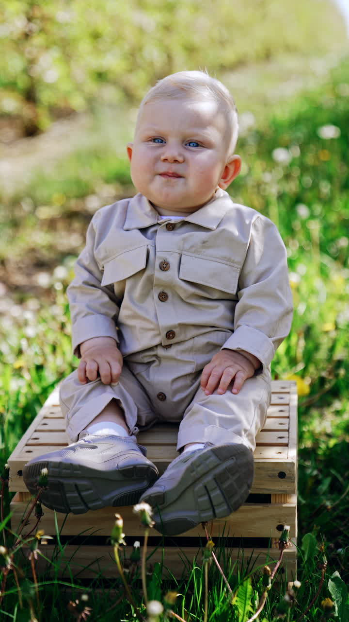 Lovely blond infant boy sitting on the crate in the spring garden. Mom's hand supports her child protecting him from falling. Vertical video.