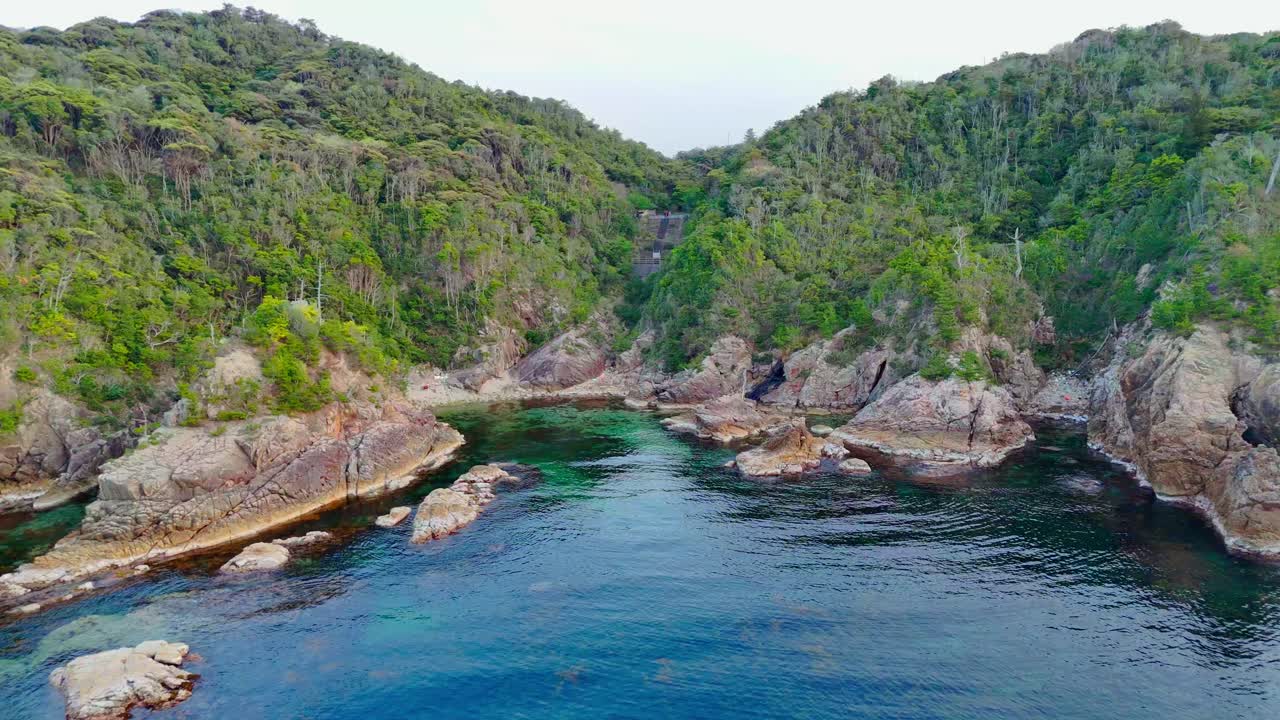 A stunning aerial pull-back shot revealing a hidden cove with clear, deep blue and turquoise water. The bay is surrounded by lush green hills and rugged, rocky cliffs, creating a pristine paradise