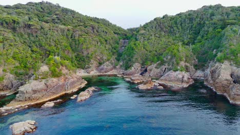 a stunning aerial pull-back shot revealing a hidden cove with clear, deep blue and turquoise water. the bay is surrounded by lush green hills and rugged, rocky cliffs, creating a pristine paradise