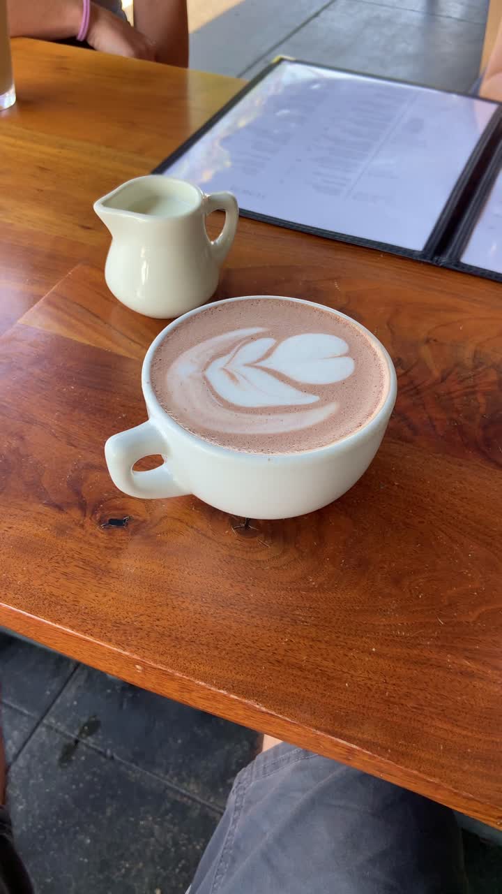 Coffee cup with leaf drawing in a restaurant or cafe shop. POV from phone or food critic content creator influencer filming table. UGC vlog style. Tasty drink in hip place. User generated content