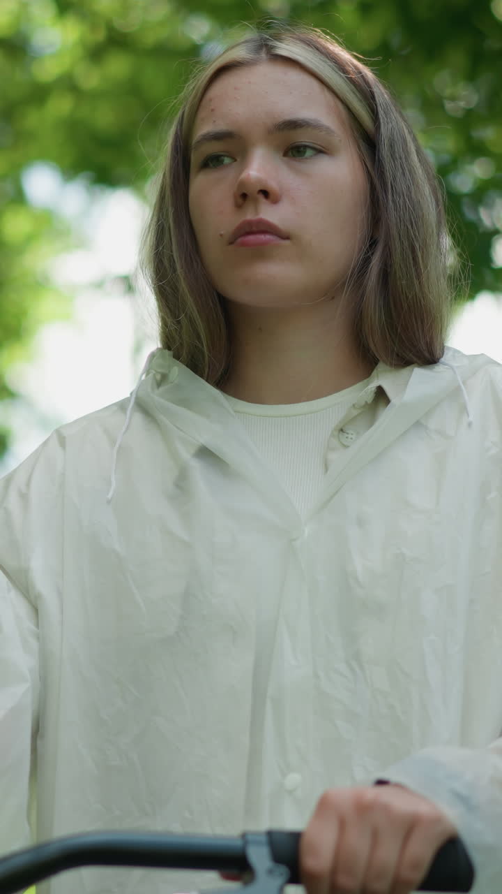 mujer en impermeable translúcido se encuentra al lado de su bicicleta, quitando la mano del manillar mientras toma una respiración profunda, mirando pensativo, rodeado de árboles verdes exuberantes, la luz del sol se filtra a través