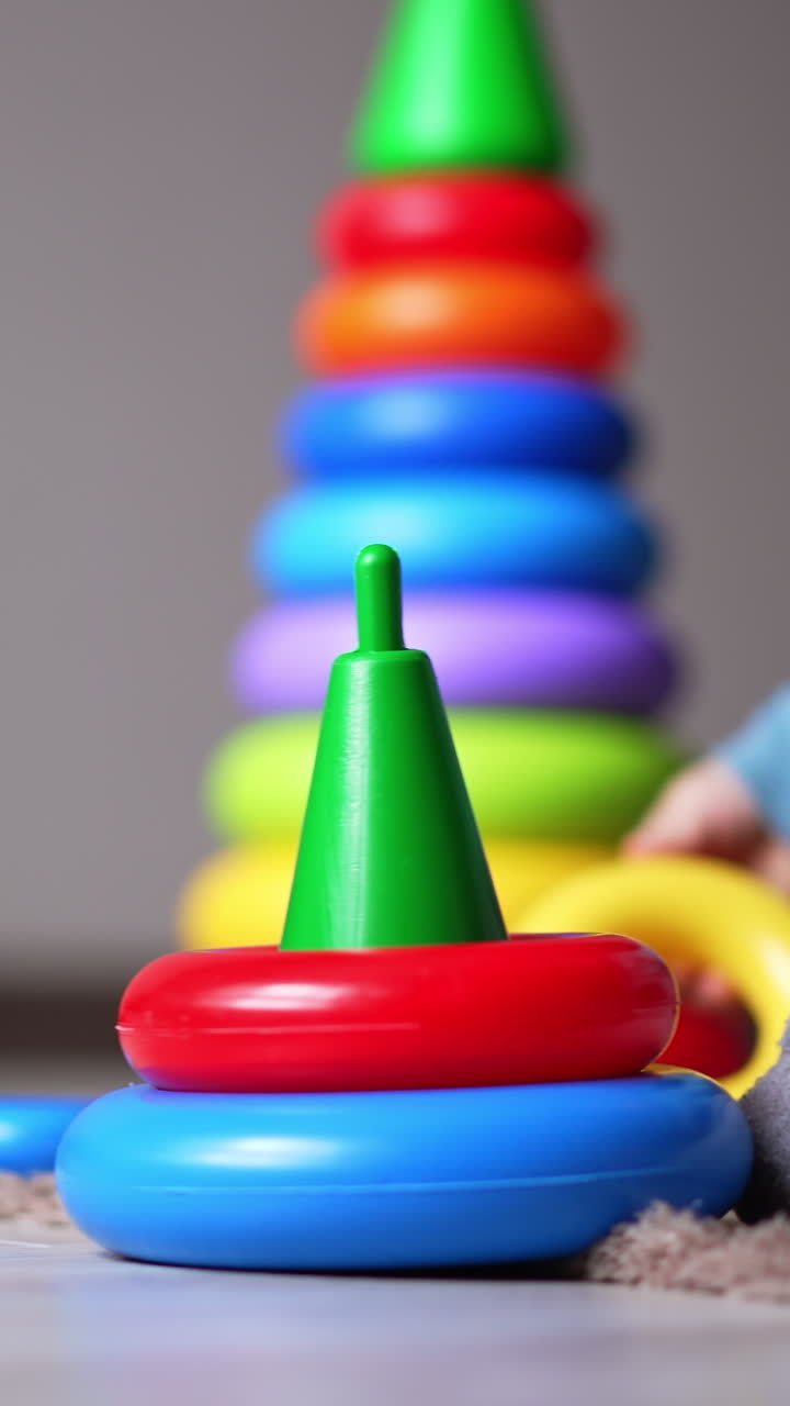 Small baby playing with little toy pyramid on the floor. Kid put the ring on and off the basis. Bigger pyramid at backdrop in blur. Vertical video