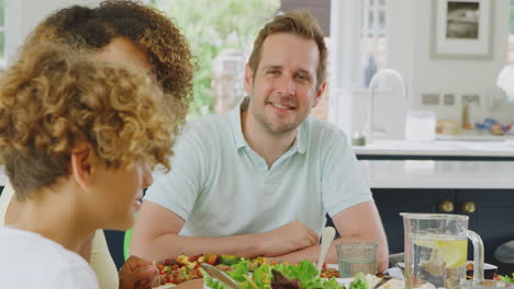 Multi-Racial-Family-Sitting-Around-Table-In-Kitchen-At-Home-Eating-Meal-Together