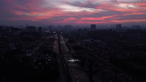 City-with-pink-sky-sunset,-golden-hour,-with-cars-passing-by-on-a-highway-aerial-drone-shot