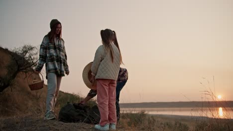 a middle-aged brunette man with gray hair in a plaid shirt collects things in a large green bag and, together with his wife and daughter, are heading home after a trip and a picnic to his city on a summer evening