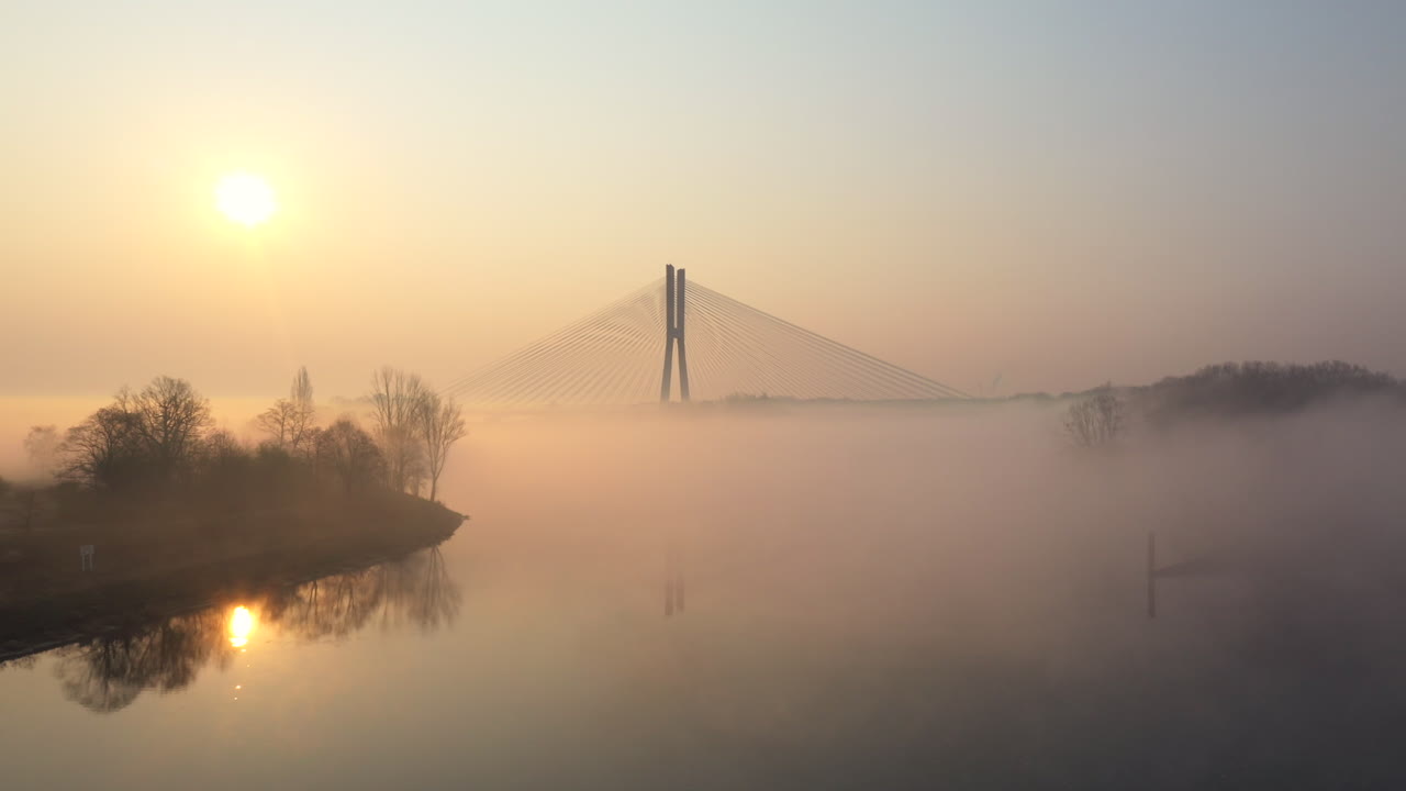 흐릿한 해가 뜨는 아침에 현대적인 케이블 다리를 향해 강을 향해 낮게 날아갑니다.