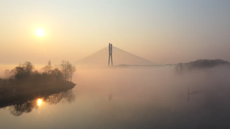 low flying towards over a river, towards a modern cable bridge during a foggy sunrise morning