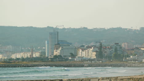 Waterfront-Buildings-In-Figueira-da-Foz,-Portugal-With-People-Swimming-In-Flowing-Water-Of-Mondego-River-In-Foreground