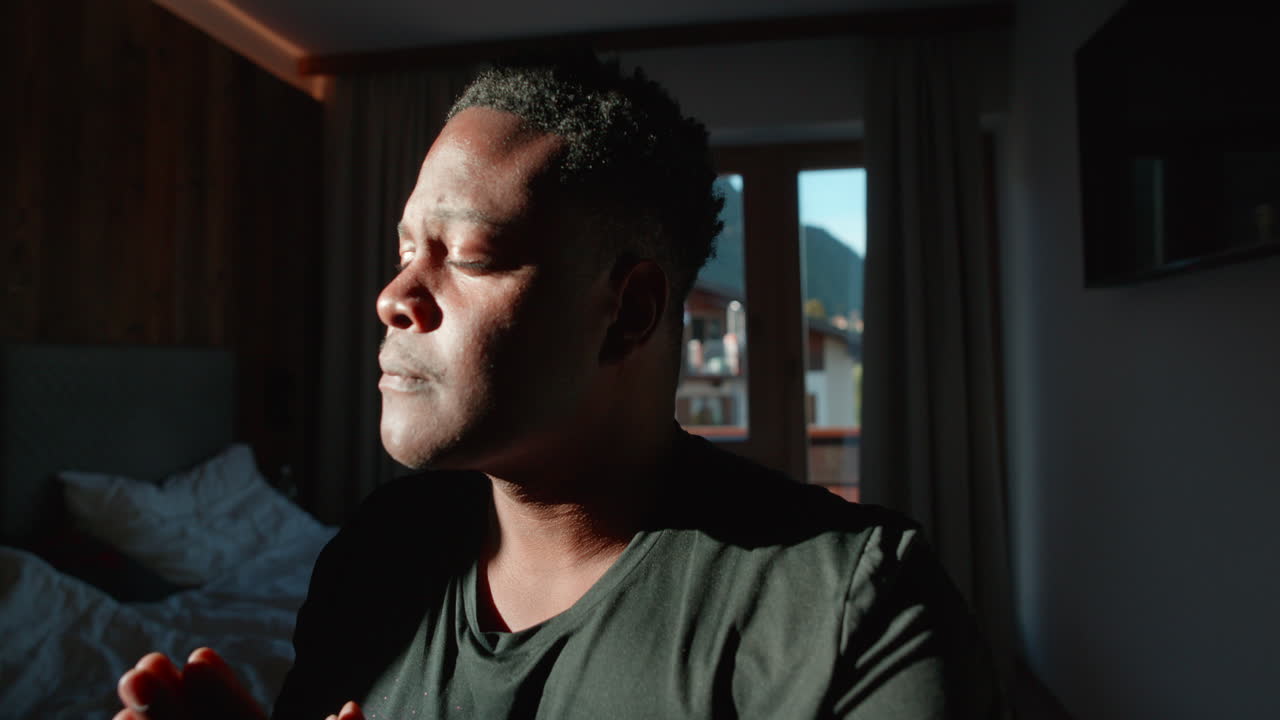 Primo piano di un uomo afroamericano solo in preghiera mattutina all'interno di una stanza buia e silenziosa.