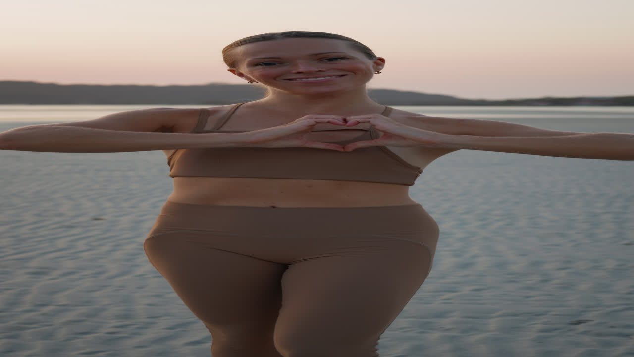 Mulher jovem formando um coração com as mãos na praia ao pôr do sol