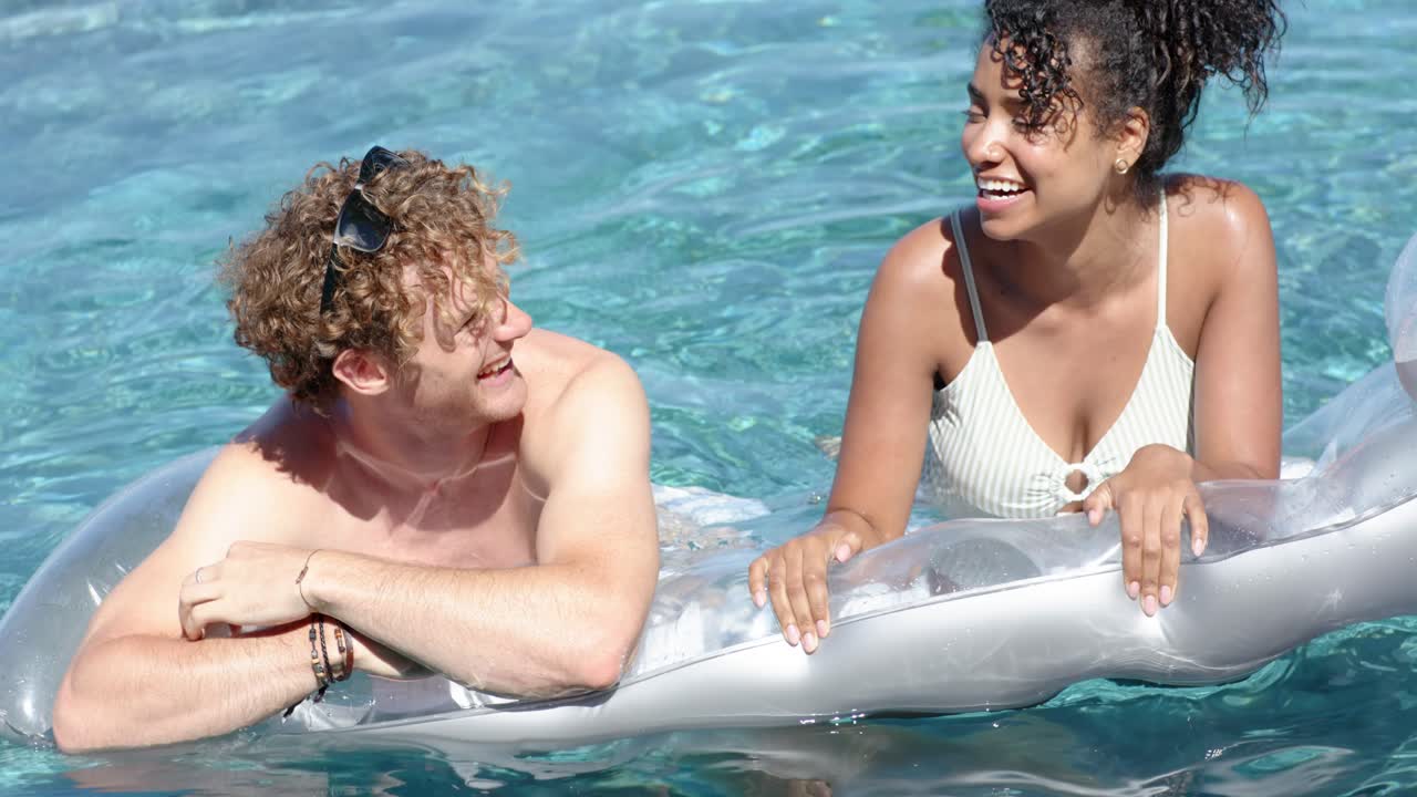 Couple relaxing on silver float in turquoise water, smiling woman touching man's shoulder flirting