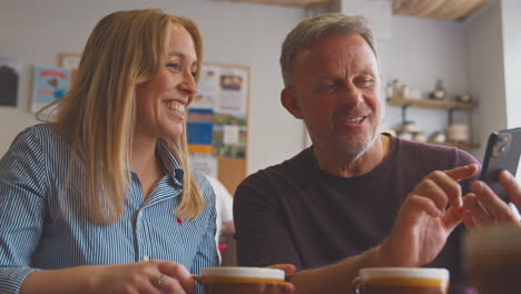 Una-Pareja-Sentada-A-La-Mesa-En-Una-Cafetería-Mirando-Juntos-El-Teléfono-Móvil