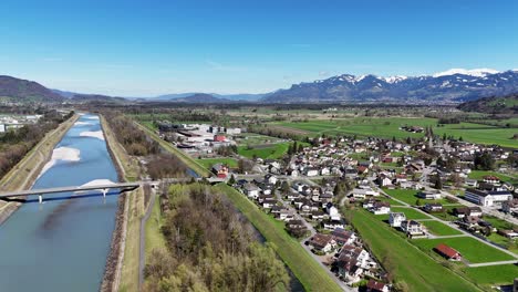 Ruggell-En-Liechtenstein-Con-Río,-Pueblo-Y-Montañas-Nevadas-Al-Fondo,-En-Un-Día-Claro,-Vista-Aérea