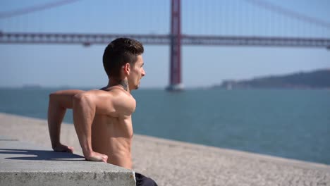 muscular shirtless tattooed man exercising at riverside