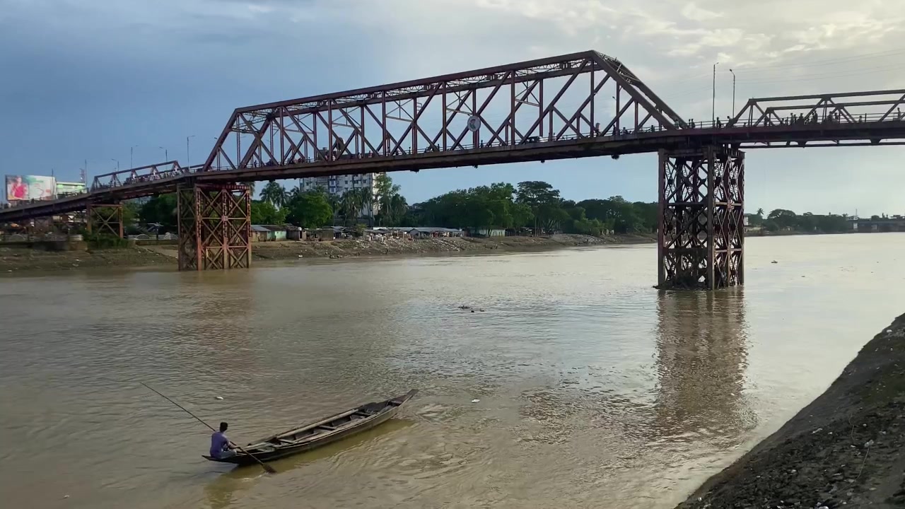 Grande ponte metallico a Sylhet, Bangladesh. Persone e traffico lo attraversano. Una barca di legno con un lungo remo si avvicina da sotto