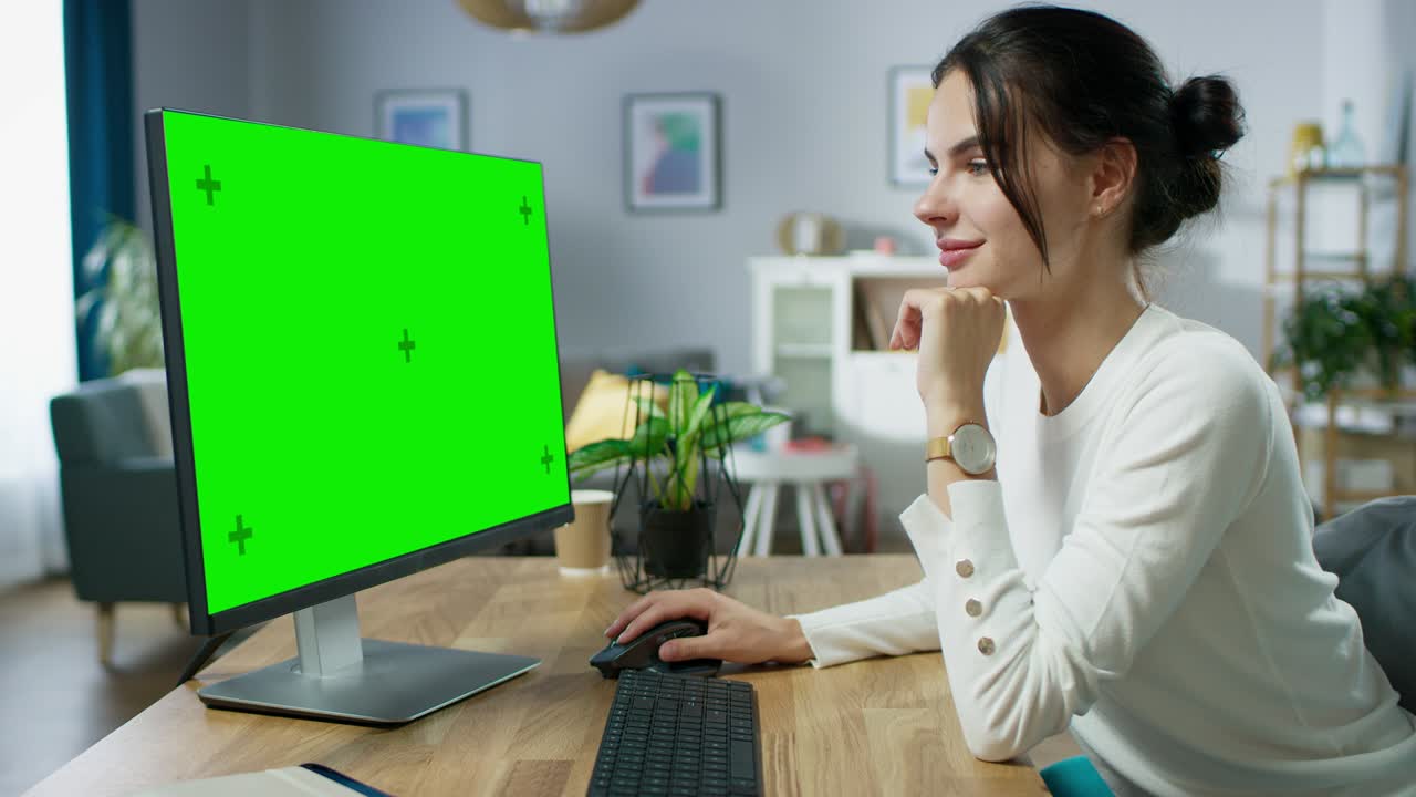 una hermosa joven trabaja en una pantalla de maqueta verde en una computadora personal mientras está sentada en su escritorio en el acogedor apartamento.