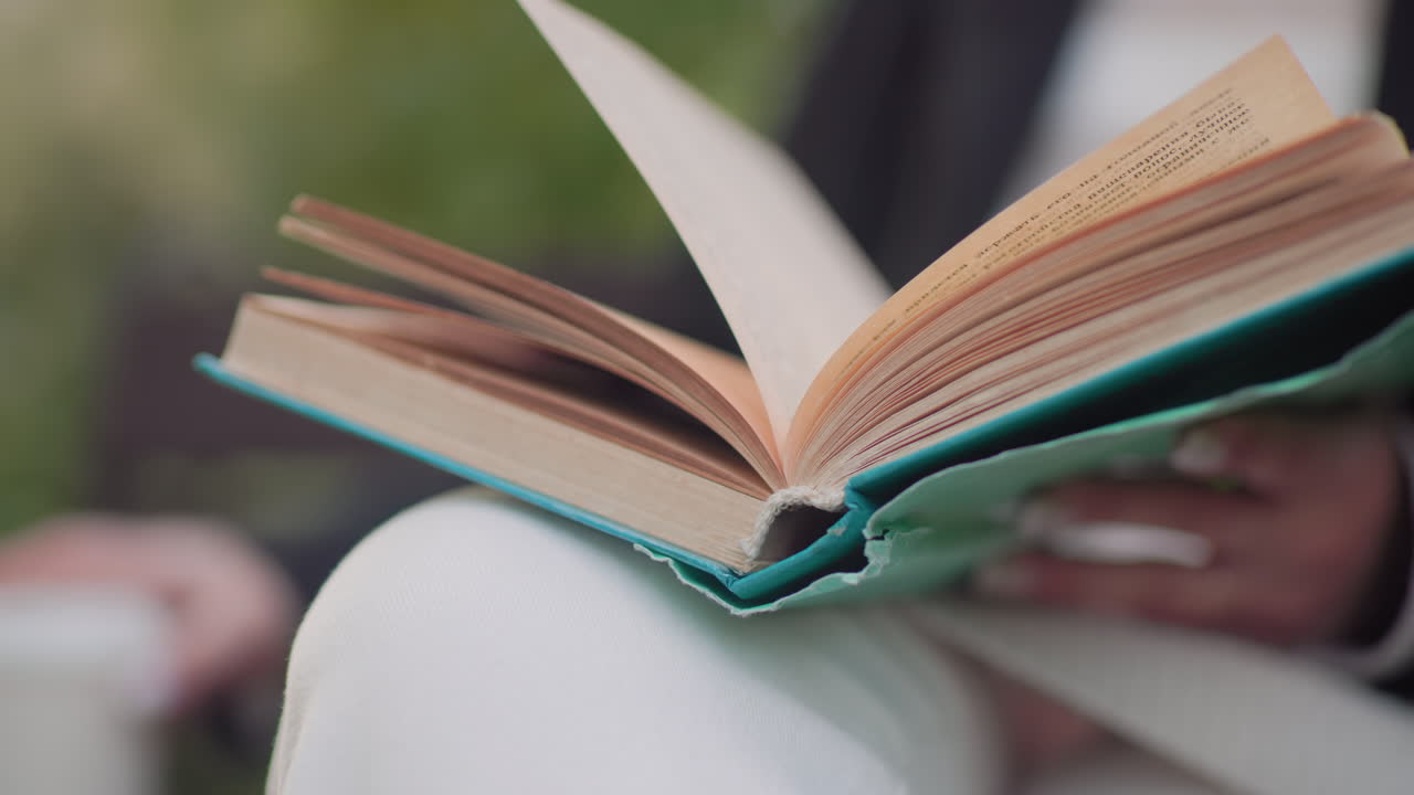 Close view of novel resting on lap of female reader with legs crossed, pages flipping in gentle breeze as hand steadies book, reaching toward nearby drink during quiet moment in green park