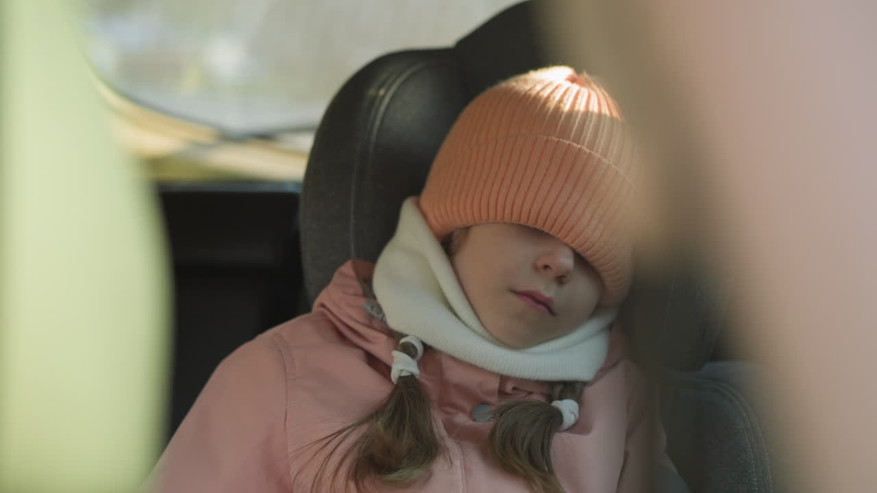 A young girl in a pink knit hat and scarf, sleeping in a car with her hat pulled down over her eyes. She appears cozy and comfortable