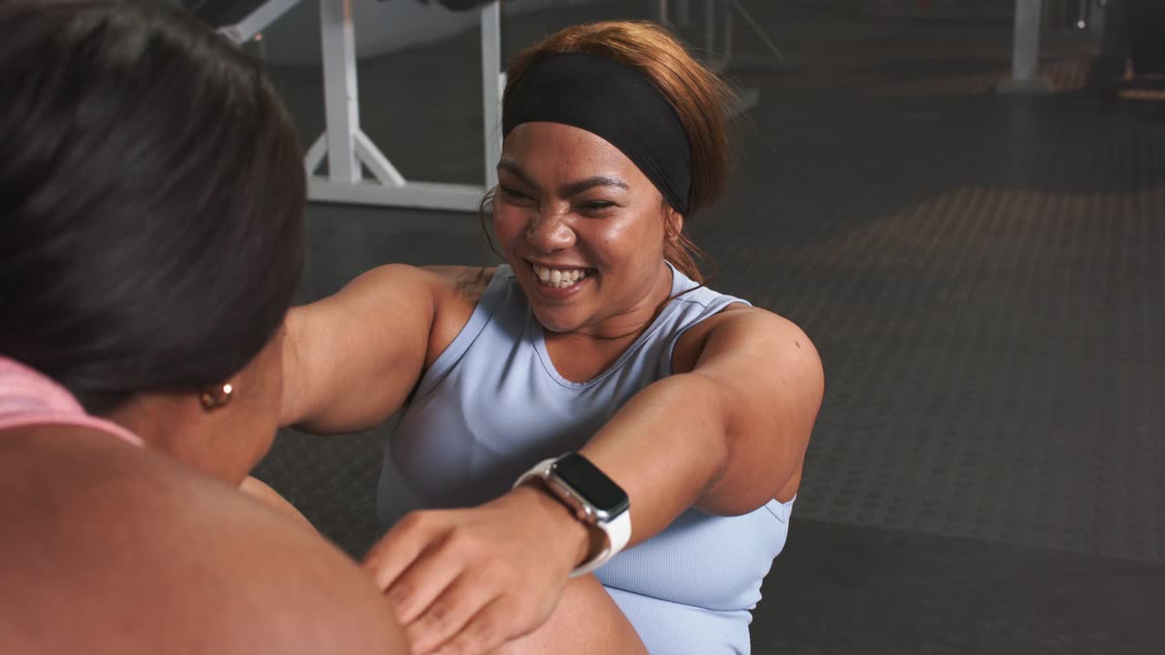 Diverse female friends grasping partner hands for sit ups crunching abs toning core on gym mat