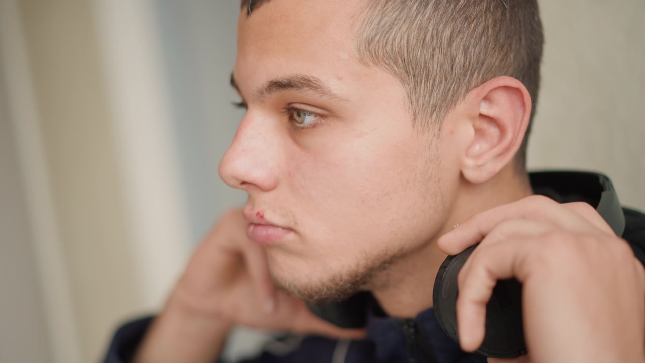 Primo piano di giovane uomo che indossa cuffie nere over-ear e le sistema con le mani. Il soggetto è mostrato di profilo su uno sfondo interno semplice, con illuminazione soffusa e toni neutri.
