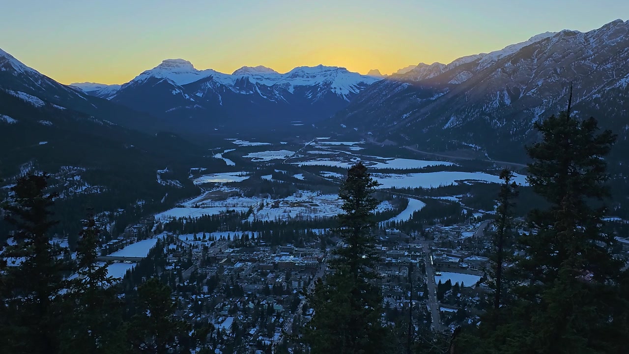 Phong cảnh mùa đông hùng vĩ ở Banff với mặt trời thấp chiếu sáng trên dãy núi Rocky của Canada, đáy thung lũng phủ tuyết, rừng cây thường xanh, các rặng núi, thị trấn đẹp như tranh vẽ bên dưới, vùng hoang dã núi cao và bầu trời trong xanh